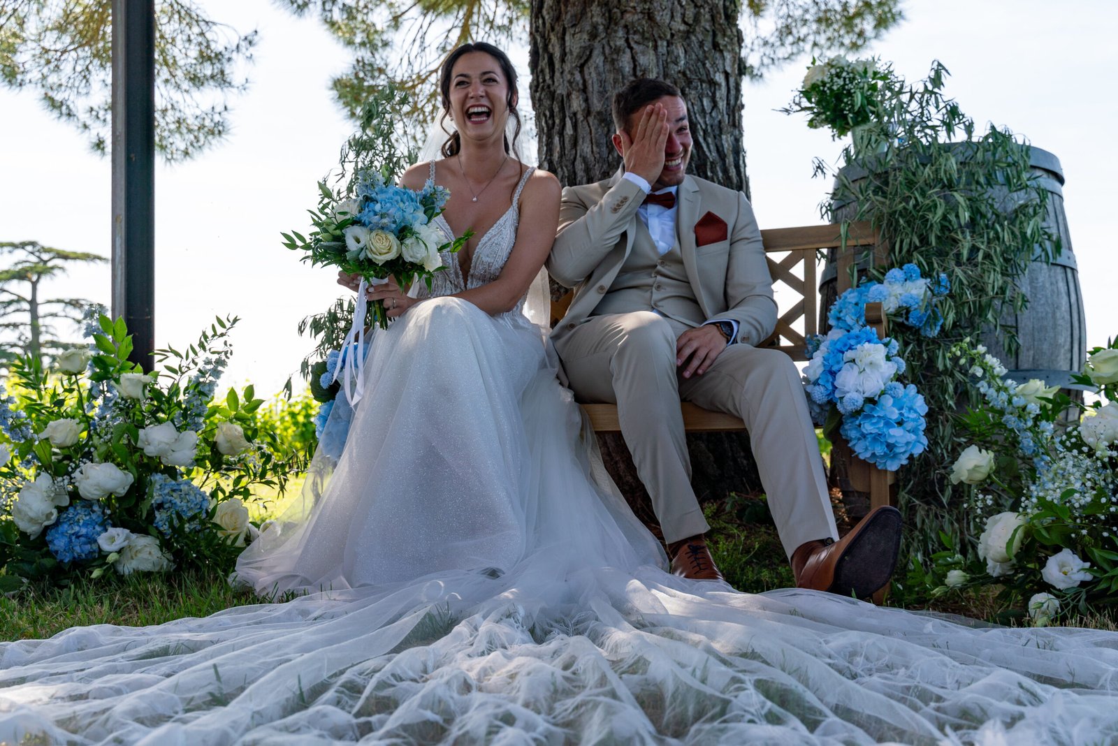 Mariés riant assis en extérieur avec bouquet de fleurs bleues, mariage à Saint-Emilion